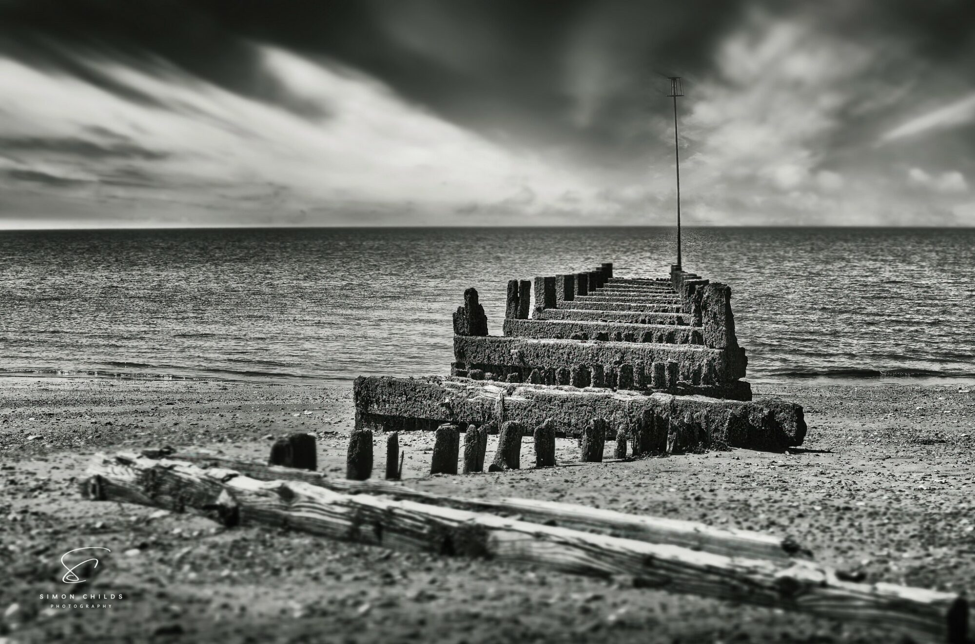 Black and white photograph of Hunstanton Beach showing dramatic skies, crashing waves, and a weathered groyne leading into the sea, captured low to the ground for enhanced depth and atmosphere.