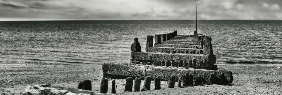 Black and white photograph of Hunstanton Beach showing dramatic skies, crashing waves, and a weathered groyne leading into the sea, captured low to the ground for enhanced depth and atmosphere.