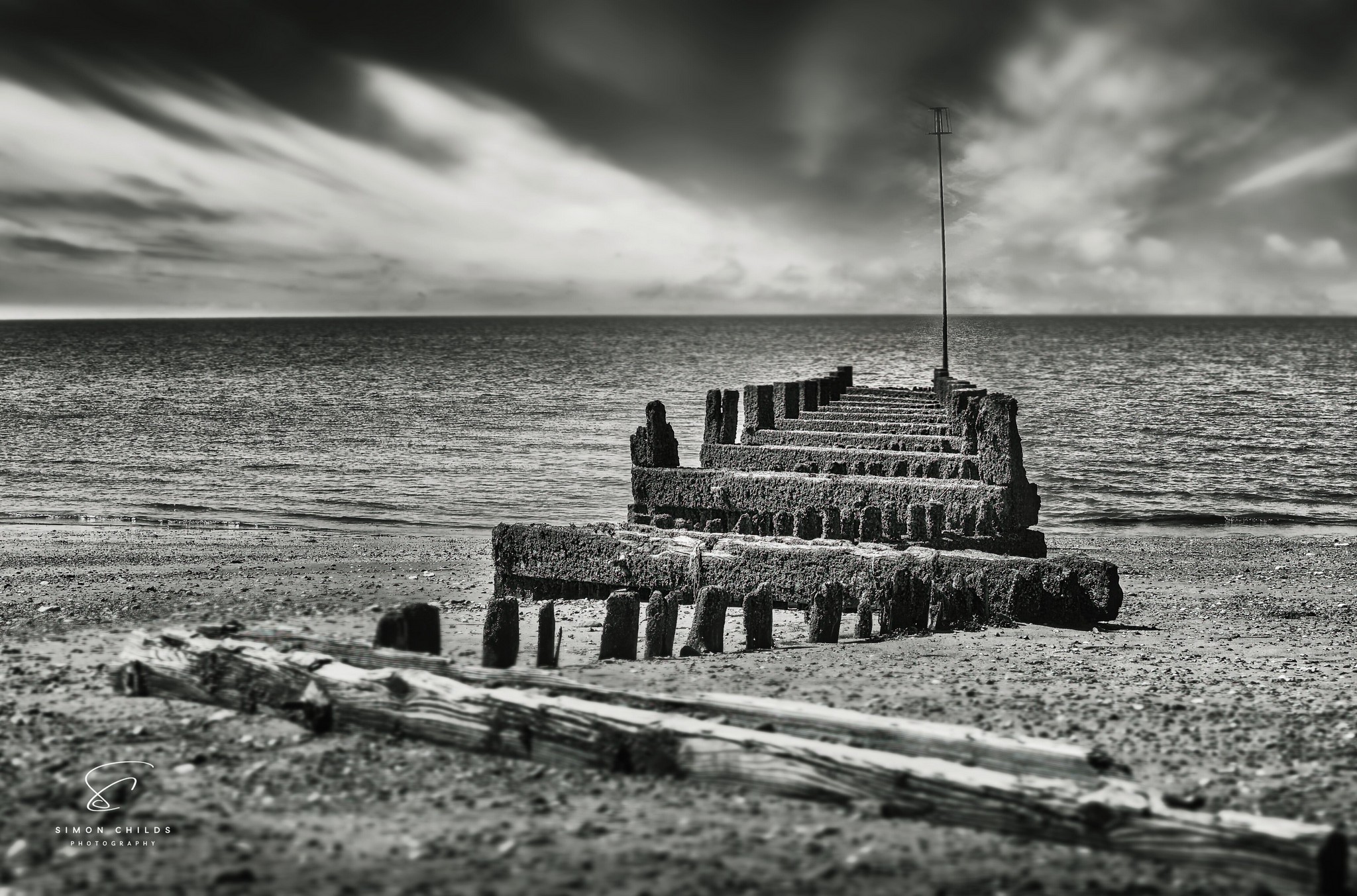 Black and white photograph of Hunstanton Beach showing dramatic skies, crashing waves, and a weathered groyne leading into the sea, captured low to the ground for enhanced depth and atmosphere.